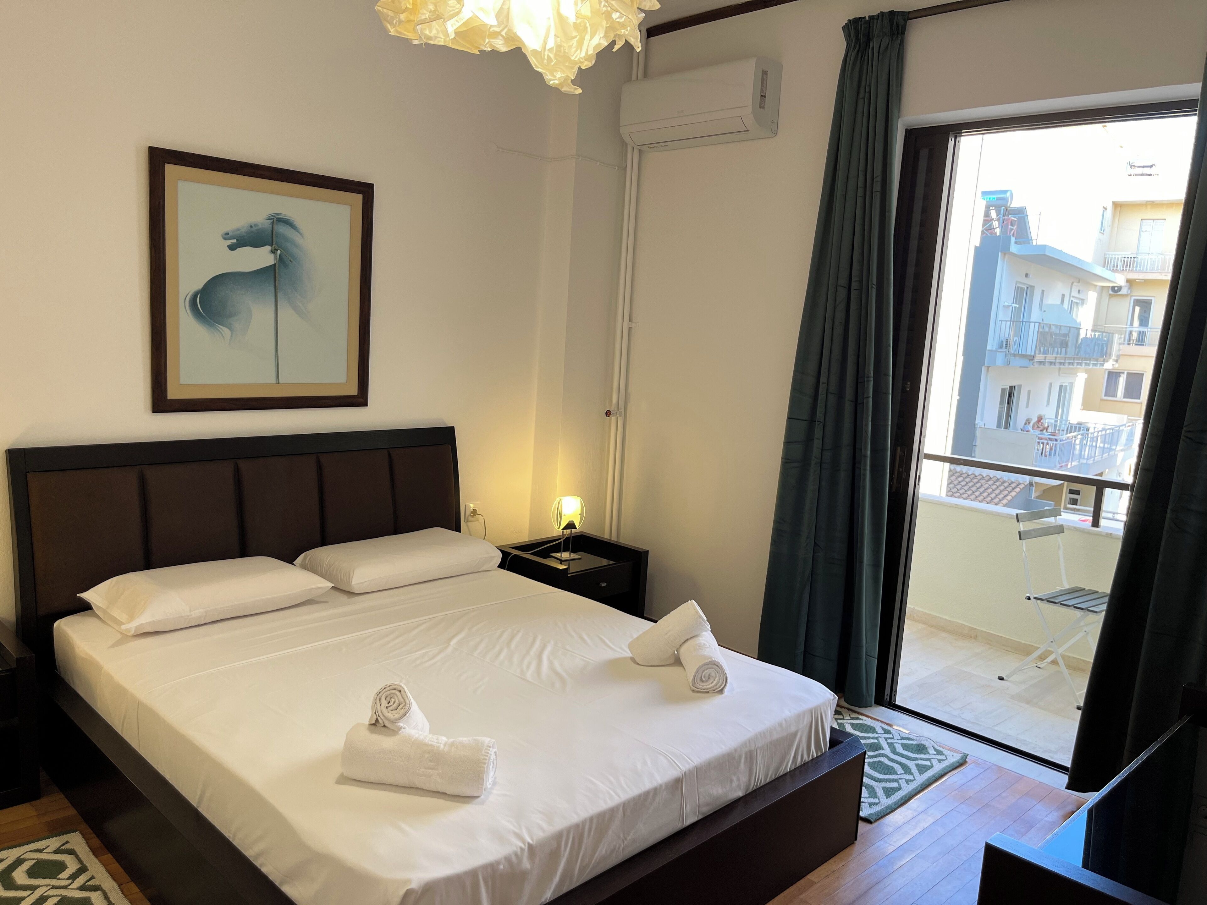 A hotel room with a large bed, a framed artwork, an air conditioning unit, a nightstand, a lamp, and a view of the outside balcony.