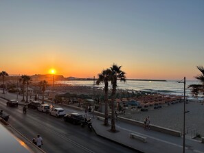 Eine Strandpromenade mit Sonnenschirmen und Sitzgelegenheiten, eine Straße mit Fahrzeugen und ein Sonnenuntergang über dem Meer.