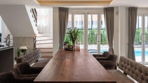 A spacious dining area with a long wooden table, cushioned chairs, and a view of a swimming pool through large windows. - Nimman villa 17 @Nimman soi17 (Chiang Mai)