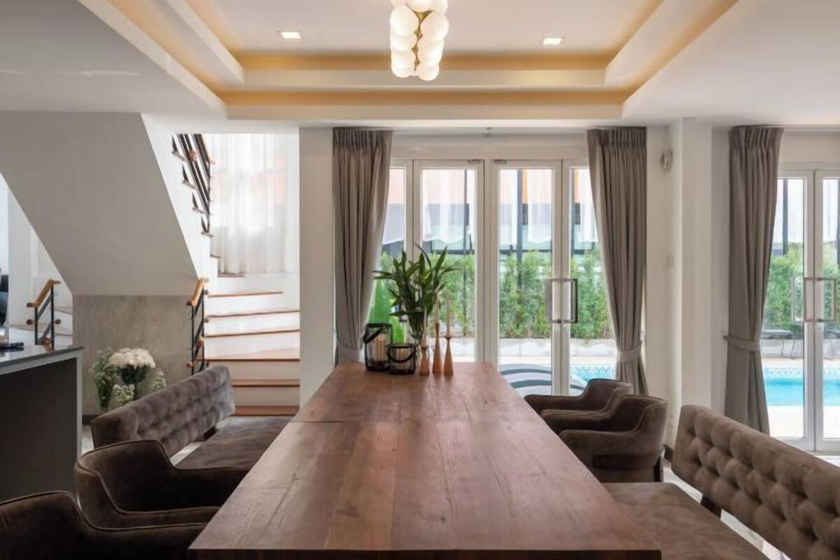 A spacious dining area with a long wooden table, cushioned chairs, and a view of a swimming pool through large windows.