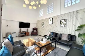 A spacious living room with a flat-screen TV, leather sofas, a coffee table, and a large window providing natural light.