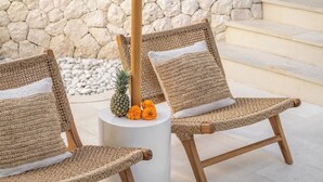 Twee rieten stoelen met witte kussens, een witte ronde tafel en een ananas met goudsbloemen erop.