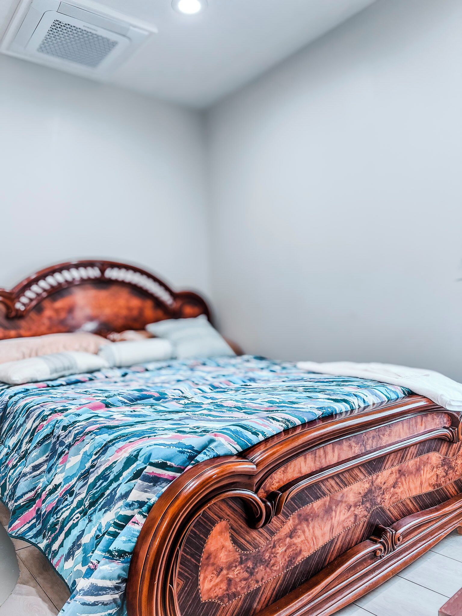 A bed with a patterned bedspread and a wooden headboard in a room with white walls and a ceiling vent.