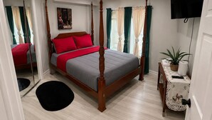 Ein Bett mit Baldachin, mit einer grau und roten Bettdecke, ein Nachttisch mit einer Pflanze und ein an der Wand befestigtes Fernsehgerät.