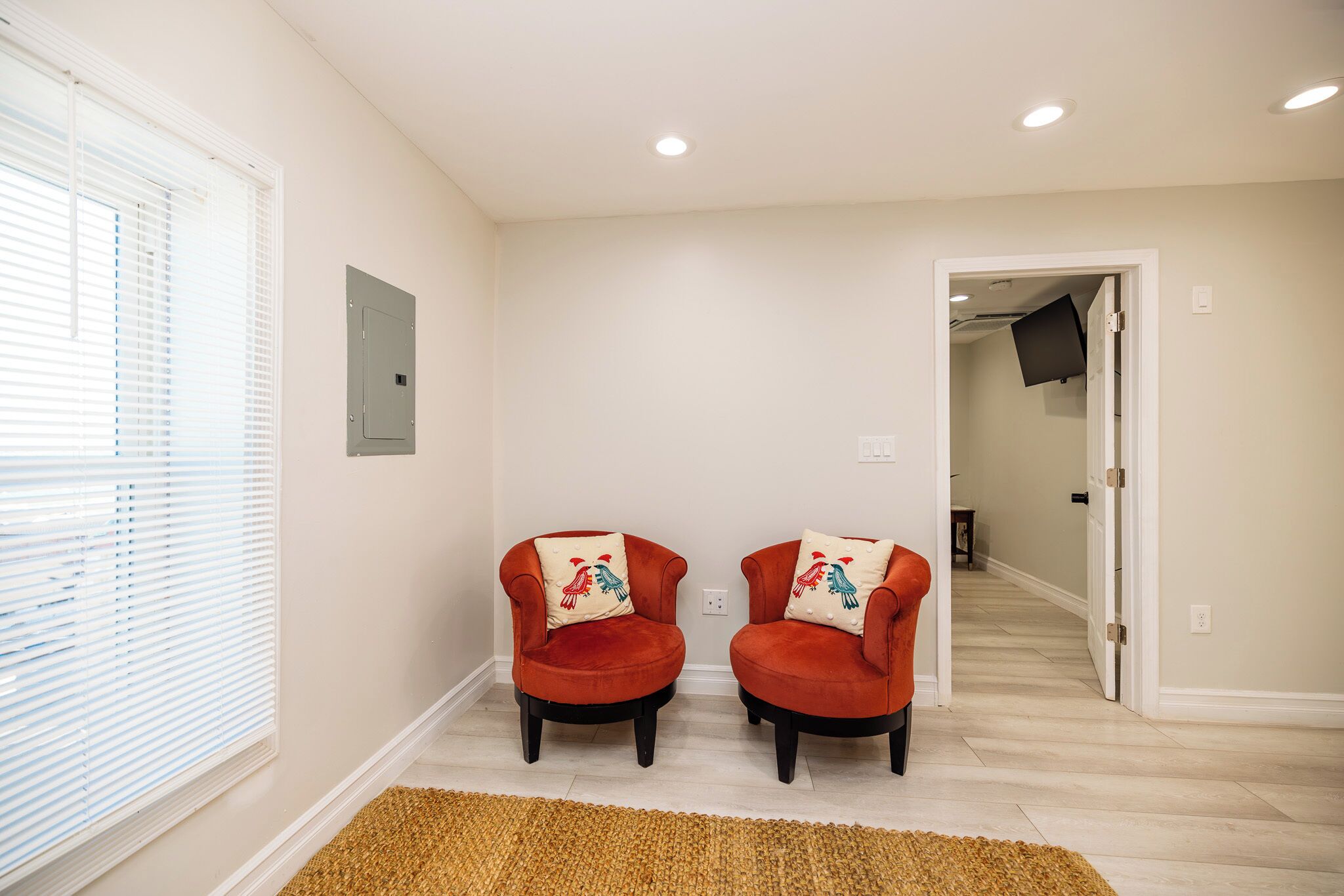 A room with two red armchairs, a wooden floor, a white wall, and a door leading to another room.