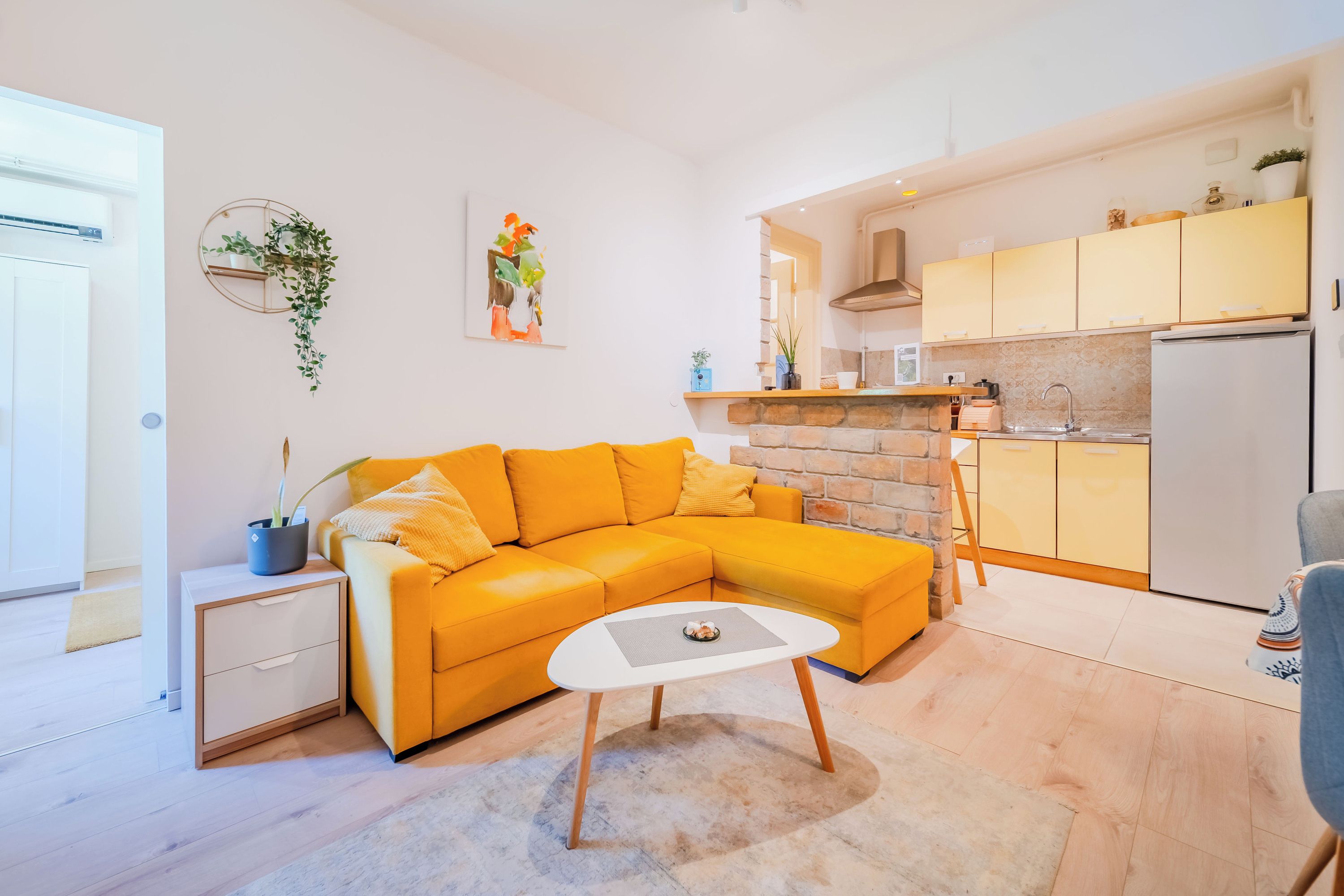 A modern living room with a yellow sofa, a small white coffee table, and a kitchen area with yellow cabinets and a stone fireplace.
