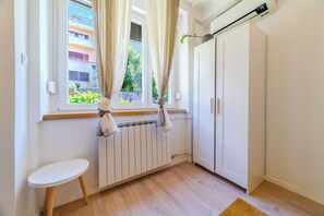 Quarto com radiador, guarda-roupa branco, mesa pequena e janela com cortinas.