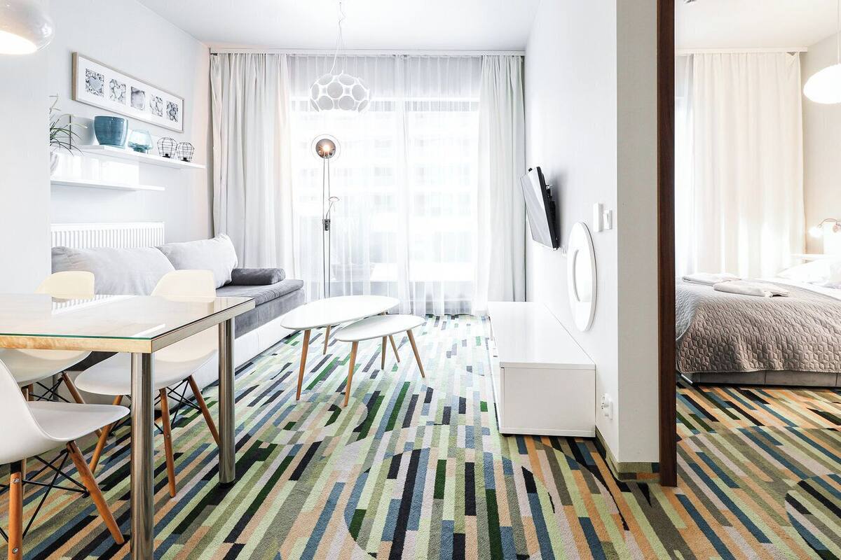 Una habitación de hotel moderna con sofá, una mesa de comedor con sillas, una pequeña cocina y un ventanal con cortinas.