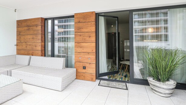 A modern living area with a sofa, a wooden panel wall, a glass door leading to a room, and a potted plant. - Baltic Park Molo by Baltic Home (Swinoujscie)
