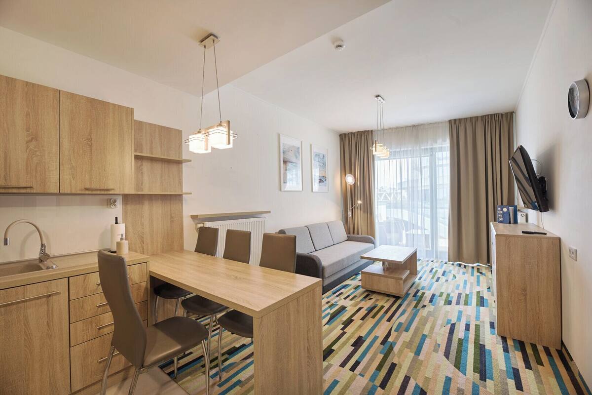 Una habitación de hotel moderna con una pequeña cocina de madera, una zona de comedor con una mesa y sillas, un sofá y un televisor sobre un mueble de madera.