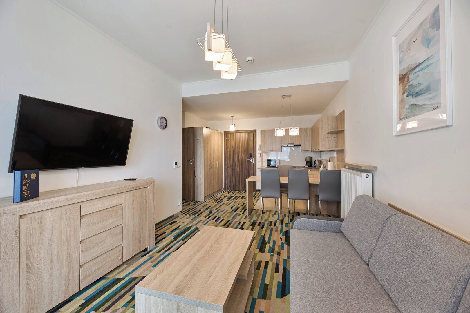 Ett modernt hotellrum med en platt-TV, en soffa, en matplats med bord och stolar samt ett litet kök.