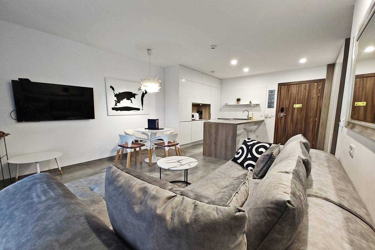 Una sala de estar moderna con un sofá gris, una mesita redonda y una cocina al fondo.