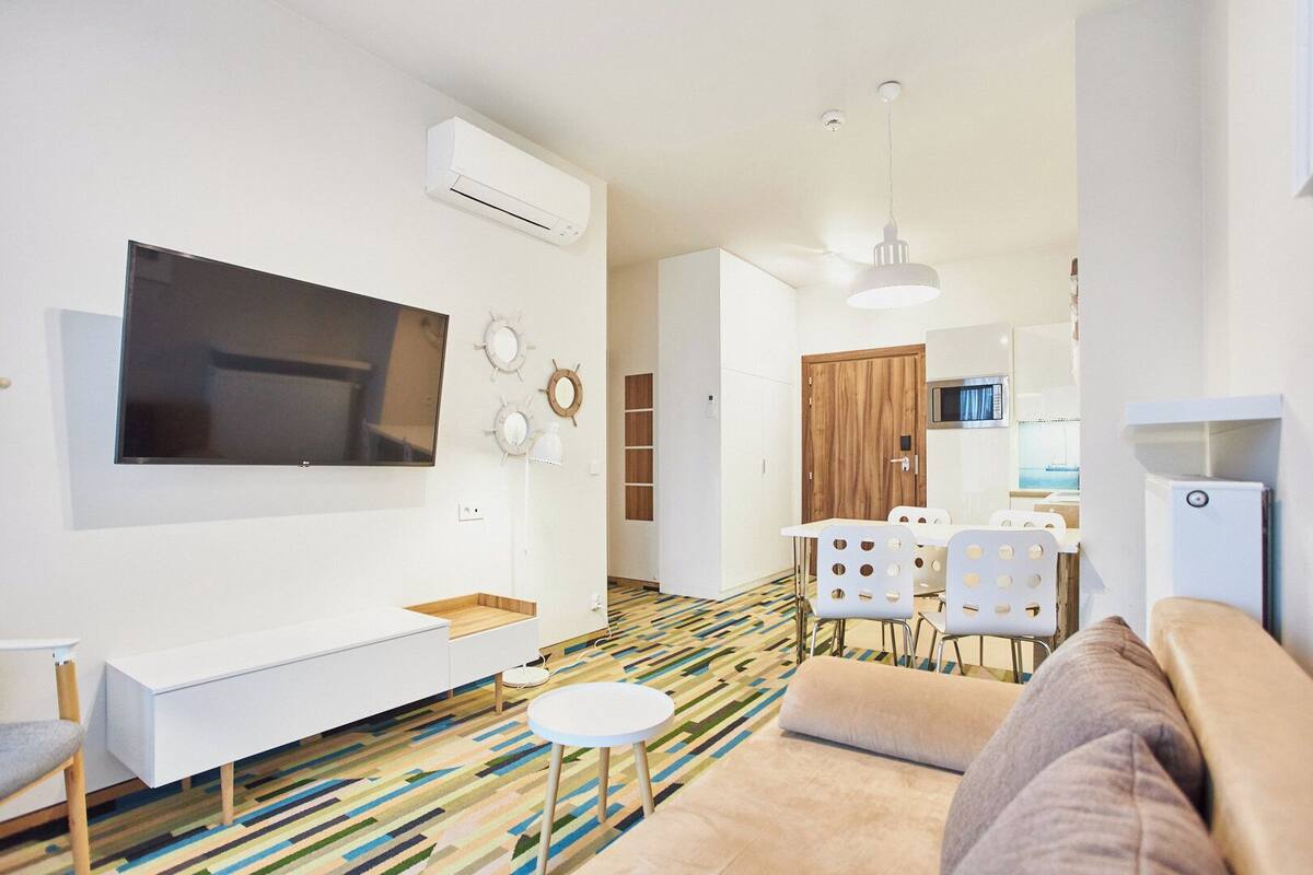 Una sala de estar moderna con televisor de pantalla plana, un sofá, un comedor con mesa y sillas, y una cocina al fondo.