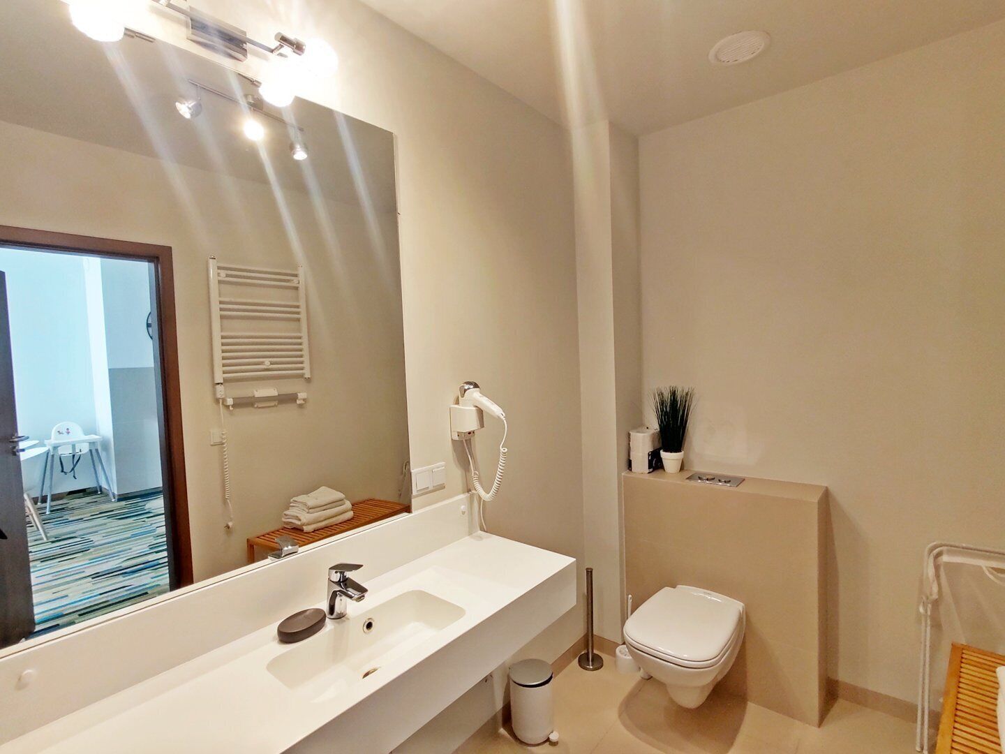 Un baño moderno con un espejo grande, un lavamanos blanco, un cabezal de ducha de mano, un inodoro y una pequeña planta en maceta.