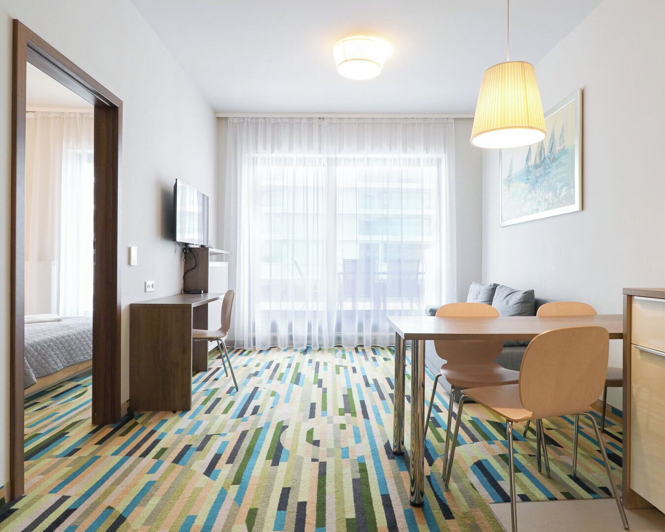 Camera d'albergo con un letto, una scrivania con sedia, un tavolo da pranzo con sedie, una TV e una finestra con tende leggere.