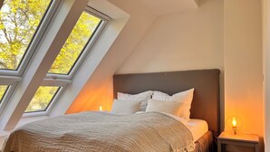 A bedroom with a sloped ceiling, a bed with white bedding, and a nightstand with a lamp. - Sommerby Sky - near the beach, shower, desk, top floor, EV charging station (Brodersby)