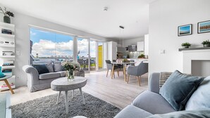 Een moderne woonkamer met een grijze bank, een salontafel en een eethoek met een tafel en stoelen. Er is een groot raam met uitzicht op een stadsgezicht.