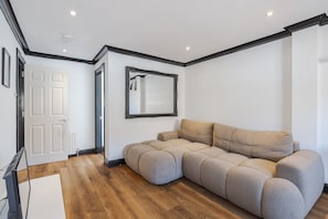 Una habitación con un sofá seccional color beige, piso de madera, una puerta blanca y un espejo enmarcado en la pared.