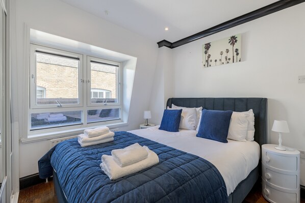 A bedroom with a large bed, a window with blinds, a nightstand, and a wall with a plant painting.