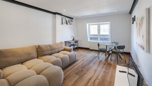 A modern living room with a sectional sofa, a dining table with chairs, a small table with a lamp, and a window with blinds. - Covent Garden’s Secret, by Veeve (London)
