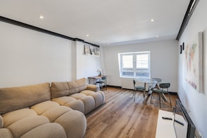 A modern living room with a sectional sofa, a dining table with chairs, a small table with a lamp, and a window with blinds. - Covent Garden’s Secret, by Veeve (London)