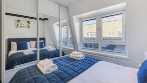 A bedroom with a large mirror, a bed with blue bedding, a window with a view of a brick building, and a radiator. - Covent Garden’s Secret, by Veeve (London)