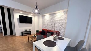 A modern living room with a white dining table, grey chairs, a red sofa, and a TV mounted on the wall.