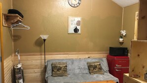 Een kleine kamer met een bed, een houten bankje, een verwarming, een klok, een klein plankje met een plant en een magnetron.