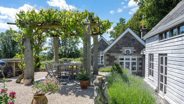 Een rustiek stenen gebouw met een houten pergola, terras en een verzorgde tuin.