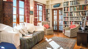Een gezellige woonkamer met een bank, fauteuils, een boekenkast en een groot raam met gordijnen.