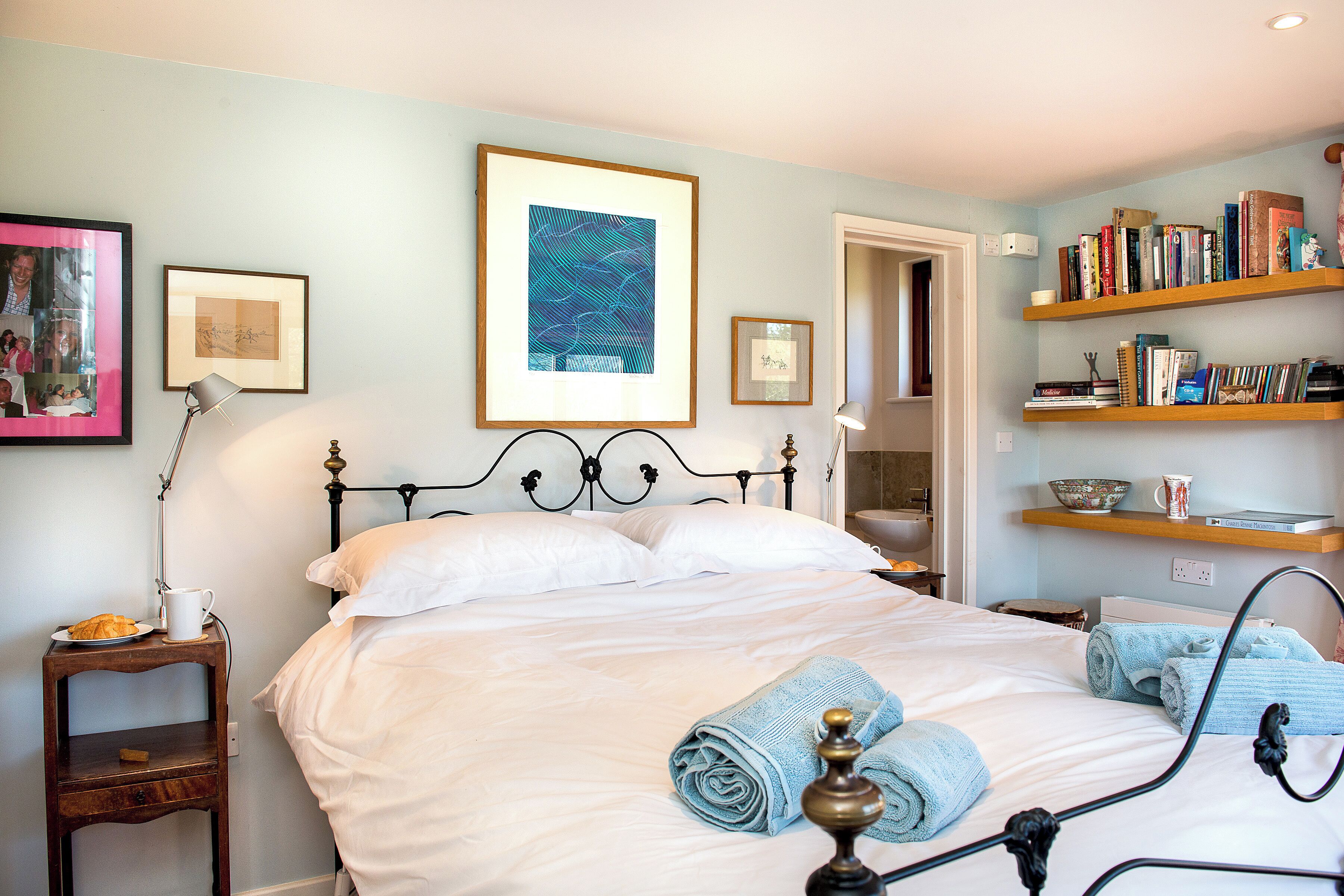 A neatly made bed with white linens, a bedside table with a lamp and a plate of croissants, and a bookshelf filled with books and decorative items.