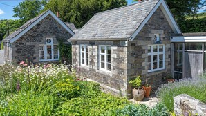 Een stenen cottage met een puntige dakschildering, omringd door een tuin met diverse planten en bloemen.