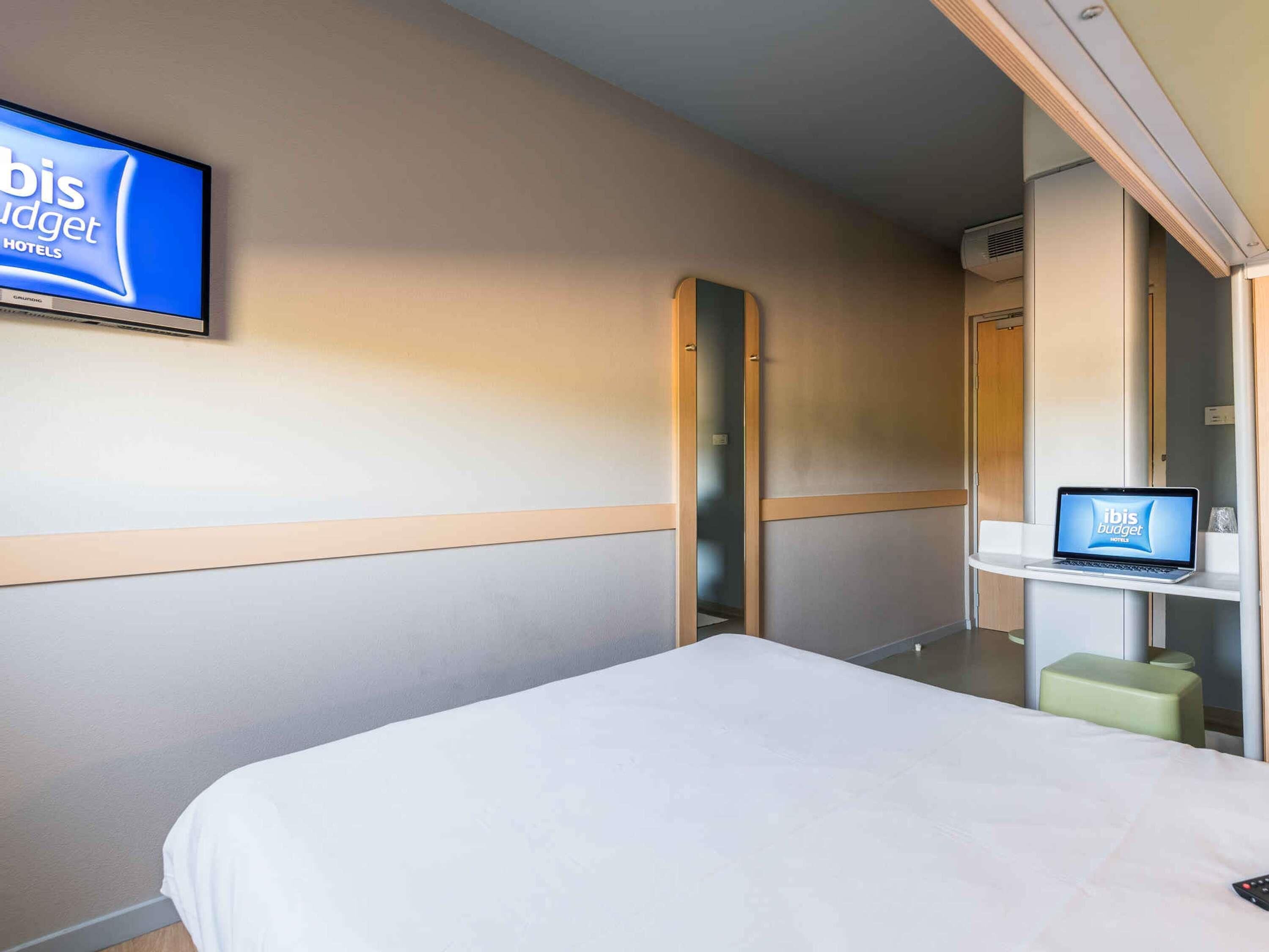 Ein Hotelzimmer mit einem Bett, einem Fernseher an der Wand, einem Laptop auf einem Regal und einem Spiegel.