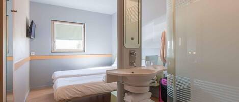 A hotel room with a bed, a sink, a mirror, a window, and a shower area.