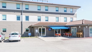 Ein zweistöckiges Hotel mit einem Schild, auf dem „ibis budget HOTEL“ steht, und einem blauen Vordach über dem Eingang. Vor dem Hotel sind zwei Autos geparkt, und ein Schild für barrierefreien Zugang ist sichtbar.