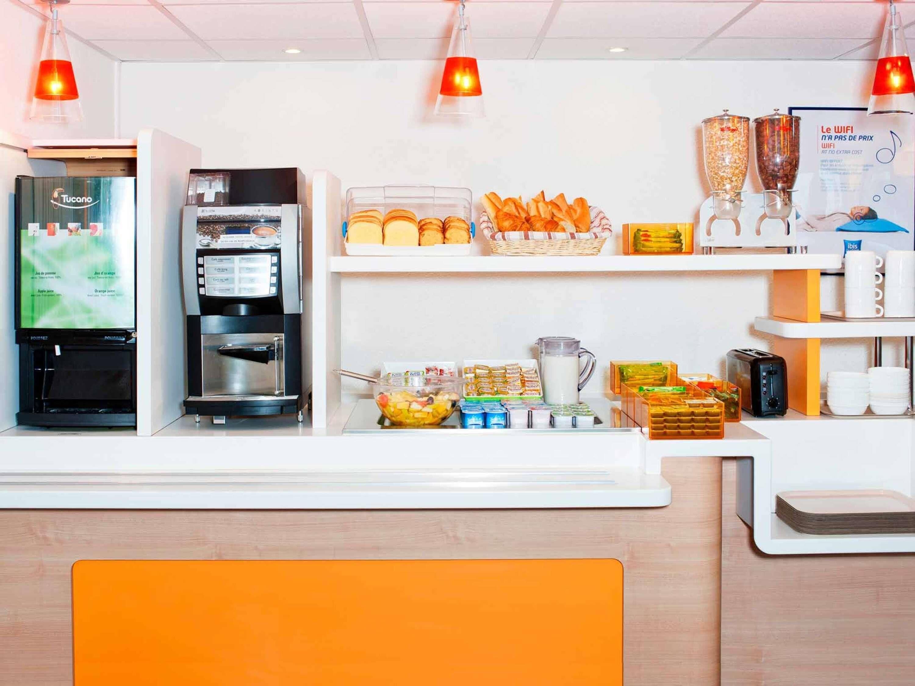Zona de recepción de hotel con una máquina de café, un refrigerador, un frutero y un bufé con pan y pastelería.