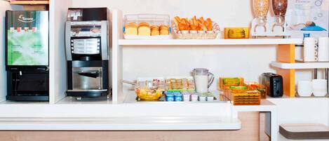 Zona de recepción de hotel con una máquina de café, un refrigerador, un frutero y un bufé con pan y pastelería.