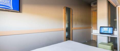 Una habitación de hotel con una cama, un televisor montado en la pared, un ordenador portátil sobre una mesita y un espejo.