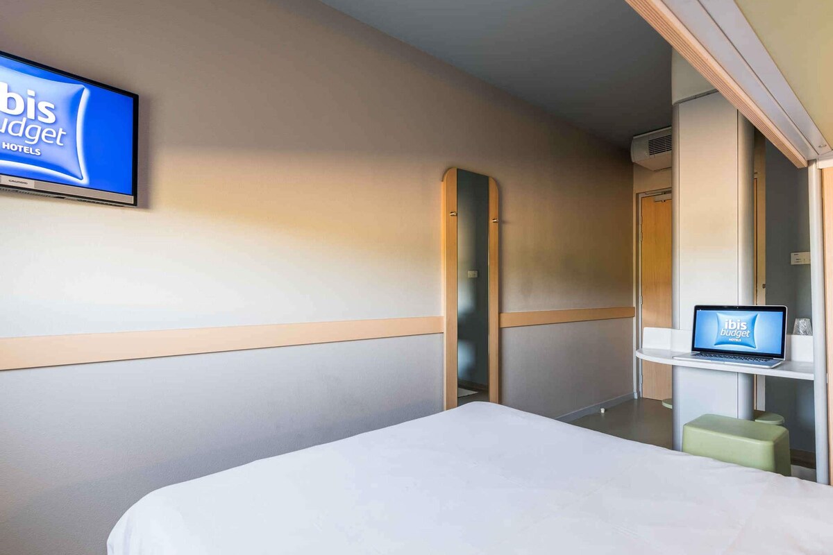 Une chambre d’hôtel avec un lit, un téléviseur fixé au mur, un ordinateur portable posé sur une étagère et un miroir.