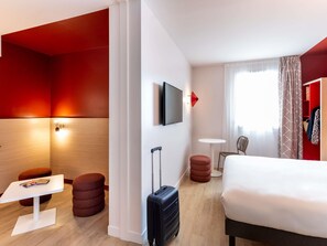 A hotel room with a bed, a small table, a chair, a red ottoman, a suitcase, a television, and a window with curtains. - ibis Styles Prevessin Geneve Airport (Prevessin-Moens)