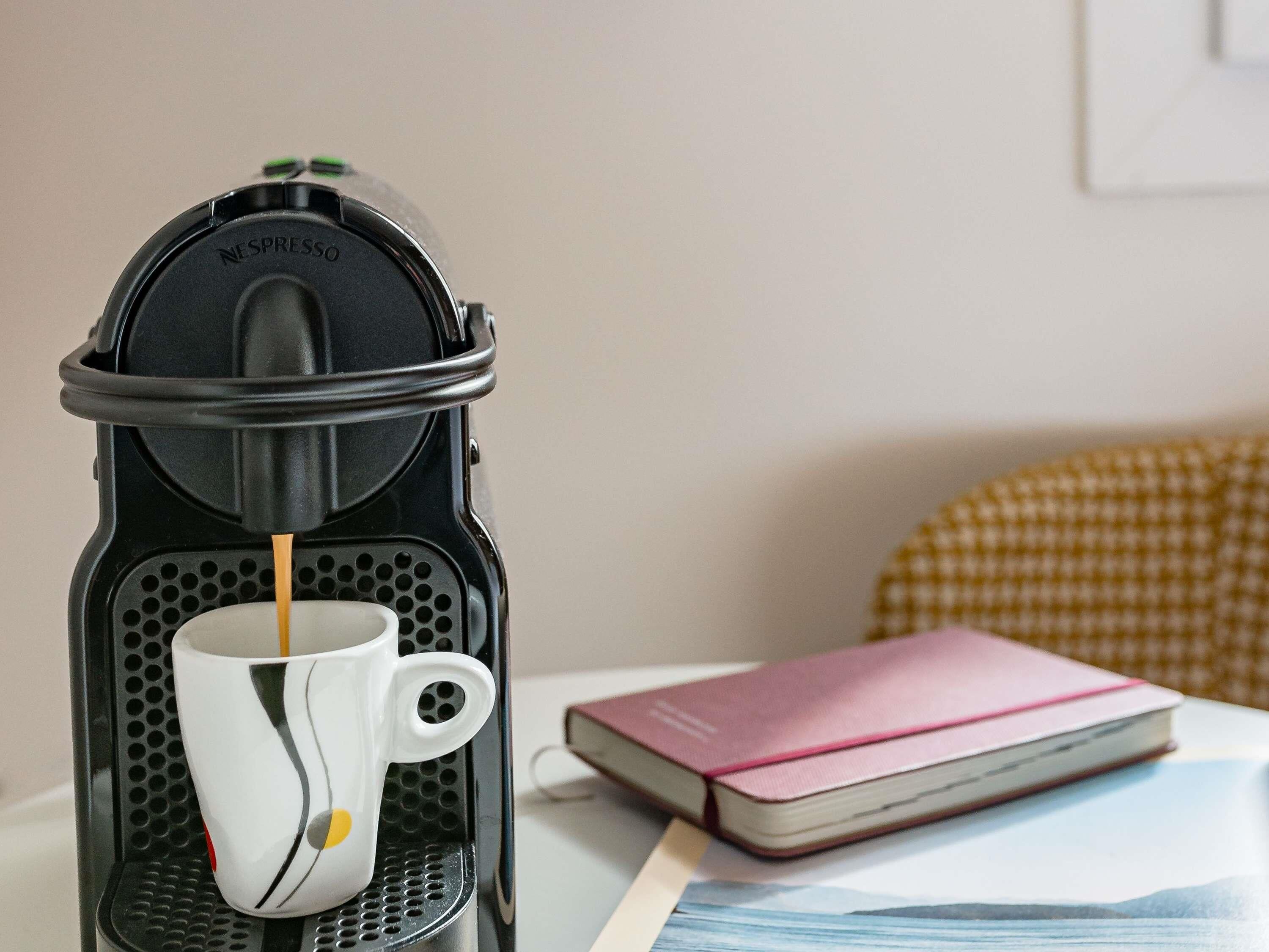 Una máquina de café Nespresso con una taza de café sirviéndose, un libro y un cojín a cuadros sobre una mesa.