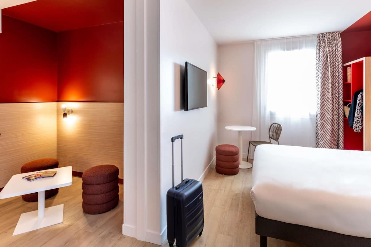 Habitación de hotel con cama, una mesita, una silla, un reposapiés rojo, una maleta, un televisor y una ventana con cortinas.