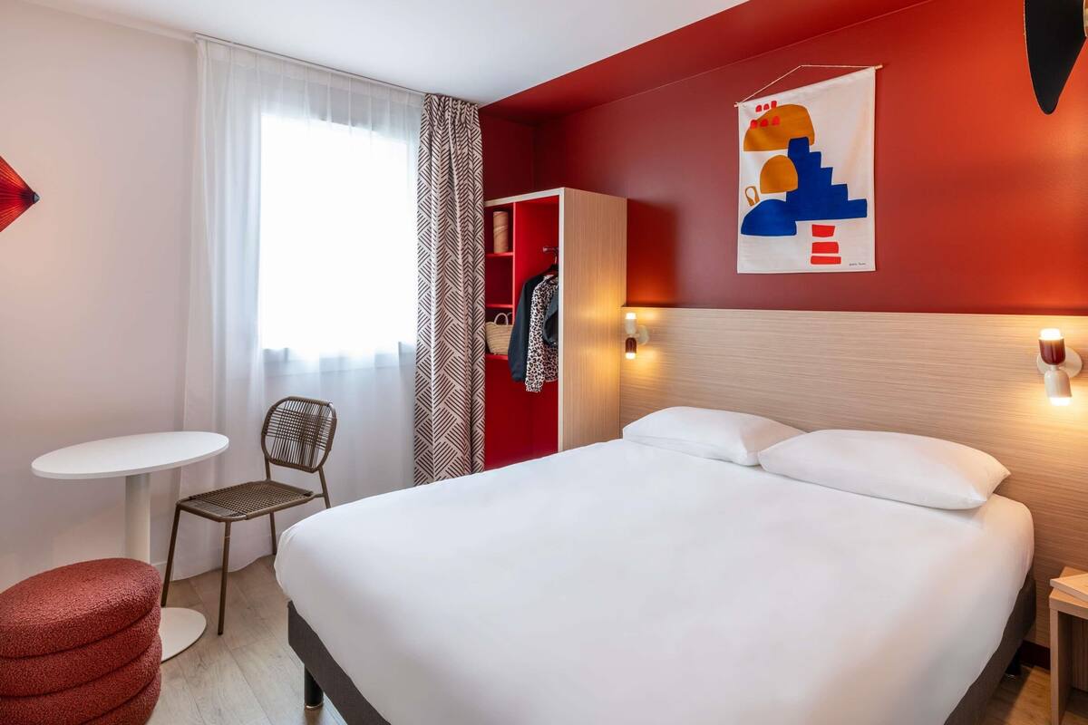 Habitación de hotel con cama, una mesita, una silla, armario y ventana con cortinas.