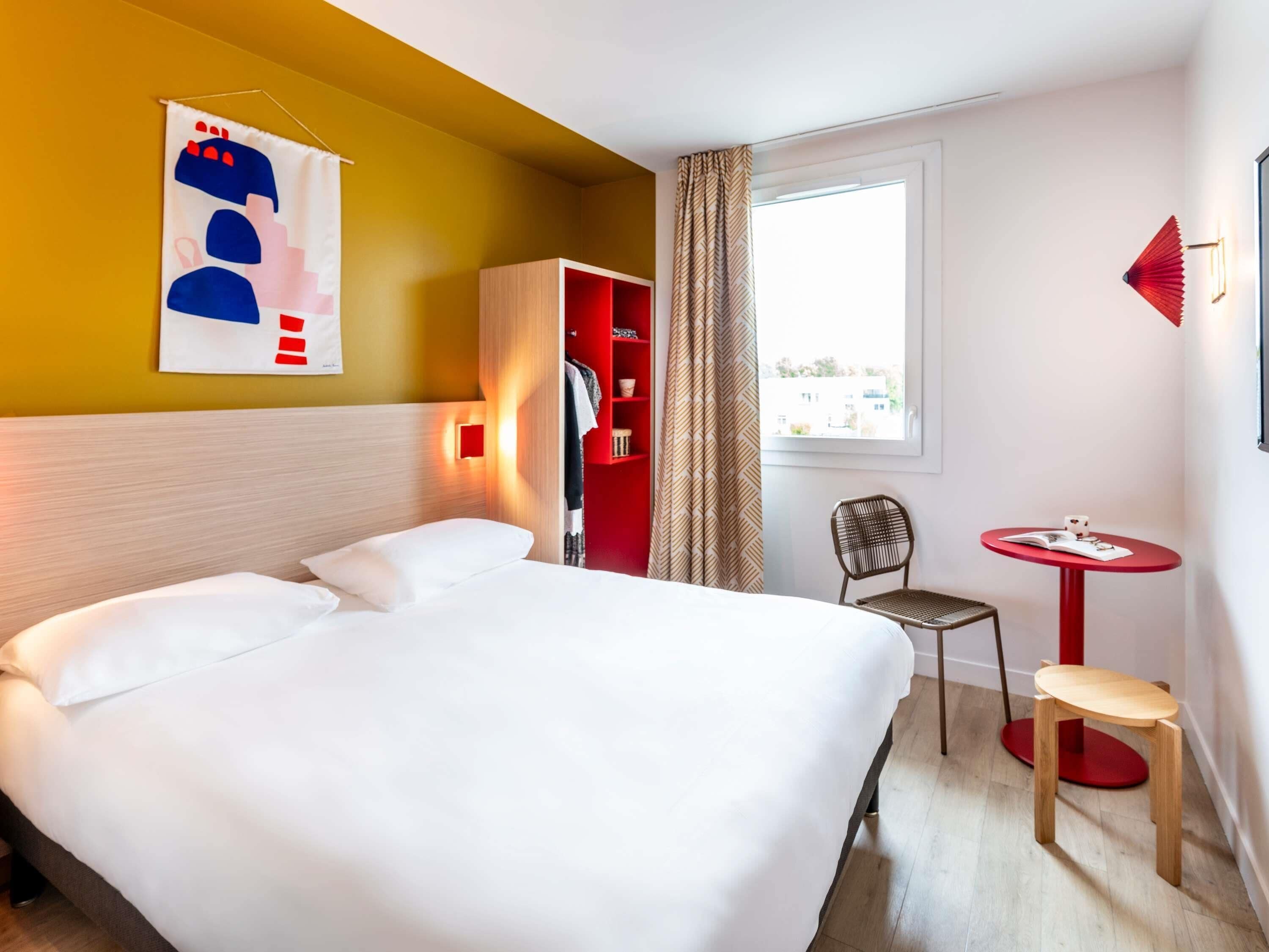 Ein Hotelzimmer mit einem großen Bett, einem roten Kleiderschrank, einem kleinen Tisch mit einem Stuhl und einem Fenster mit Blick auf Gebäude.