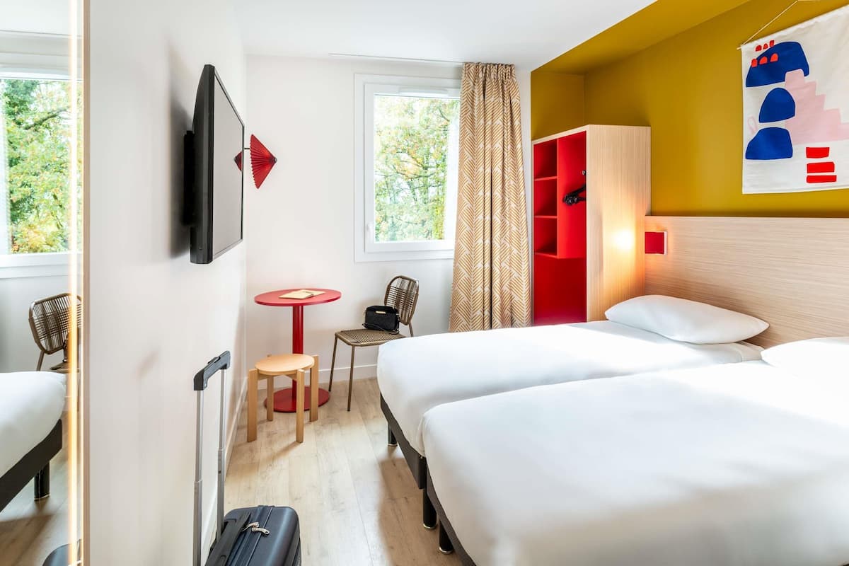 Habitación de hotel con dos camas, televisión, una mesita con una silla, un armario rojo y una ventana con cortinas.