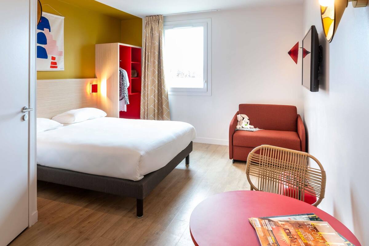 Una habitación de hotel con una cama, un sofá rojo, una silla de mimbre, una mesita y una ventana con cortinas.