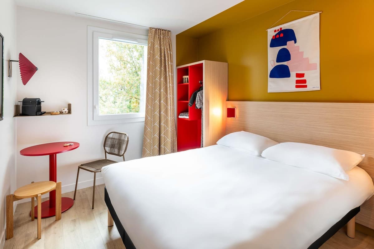 Una habitación de hotel con una cama, una mesa roja, una silla, un ventanal con cortinas y una pared con un cuadro.