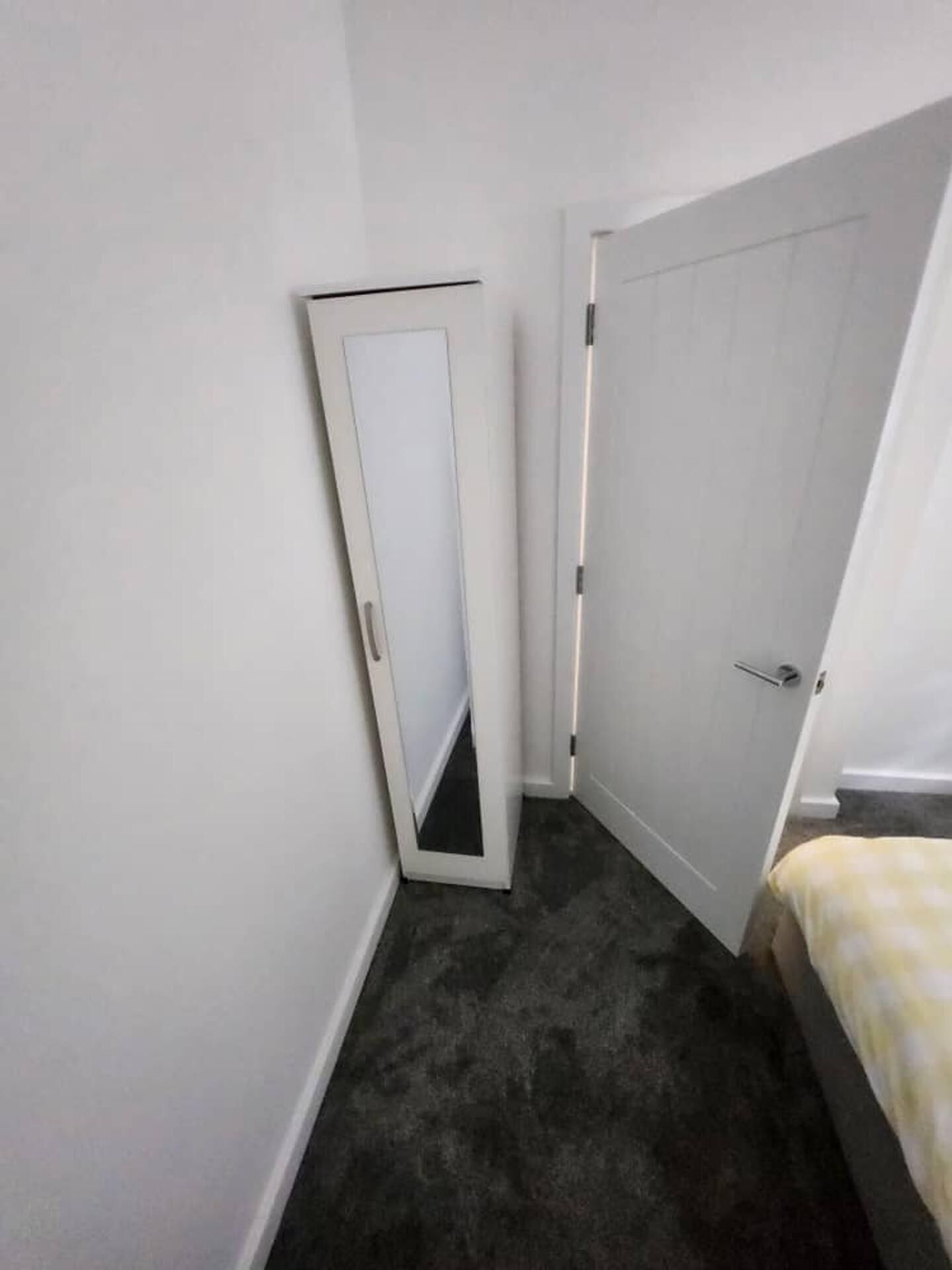Una habitación estrecha con alfombra oscura, una puerta blanca y una cama con colcha de diseño amarillo y blanco.