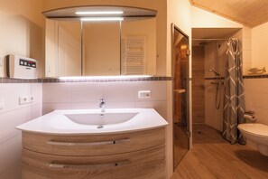 Een moderne badkamer met een witte wastafel, een houten opbergmeubel, een spiegel met ingebouwde verlichting, een douche en een toilet.