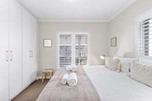 A bedroom with a bed, two rolled towels, a nightstand, a lamp, and a window with blinds. - Mayfair 246 (Cape Town)